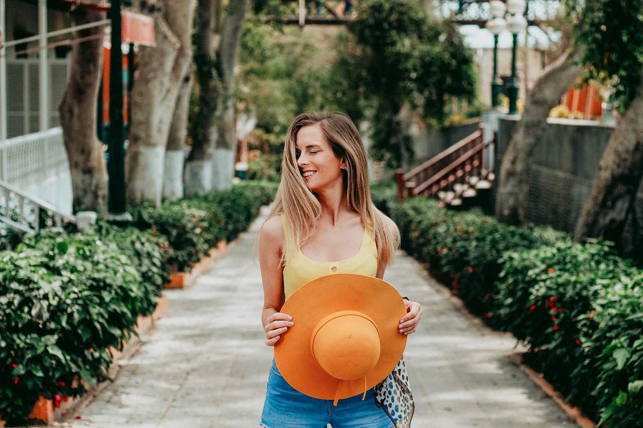 A smiling woman in a yellow top holding a sun hat, enjoying an outdoor walk, promoting sun protection and a healthy lifestyle for skin care.