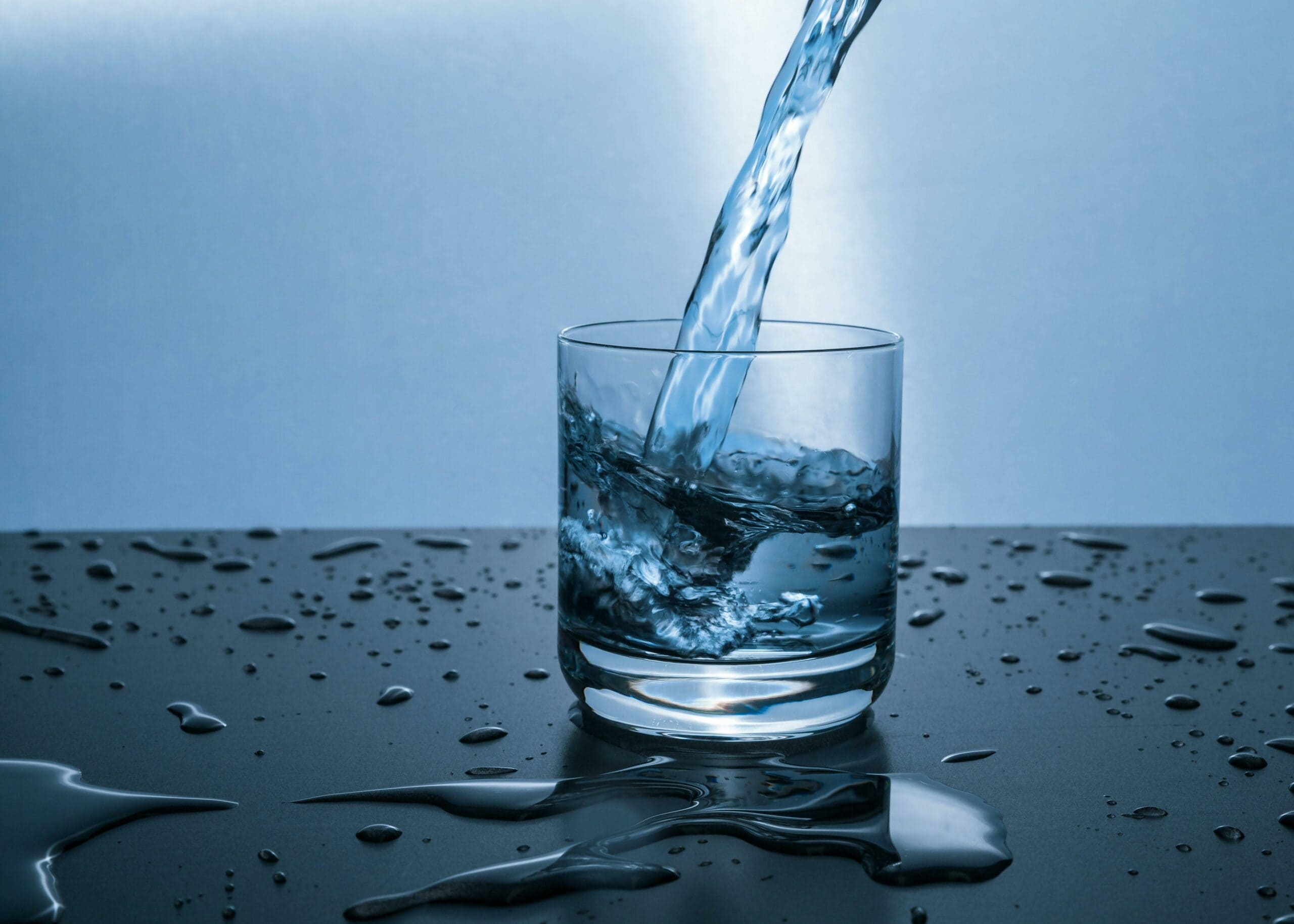 A glass of water being poured, with water droplets on the surface, symbolizing hydration and its importance for skin health.