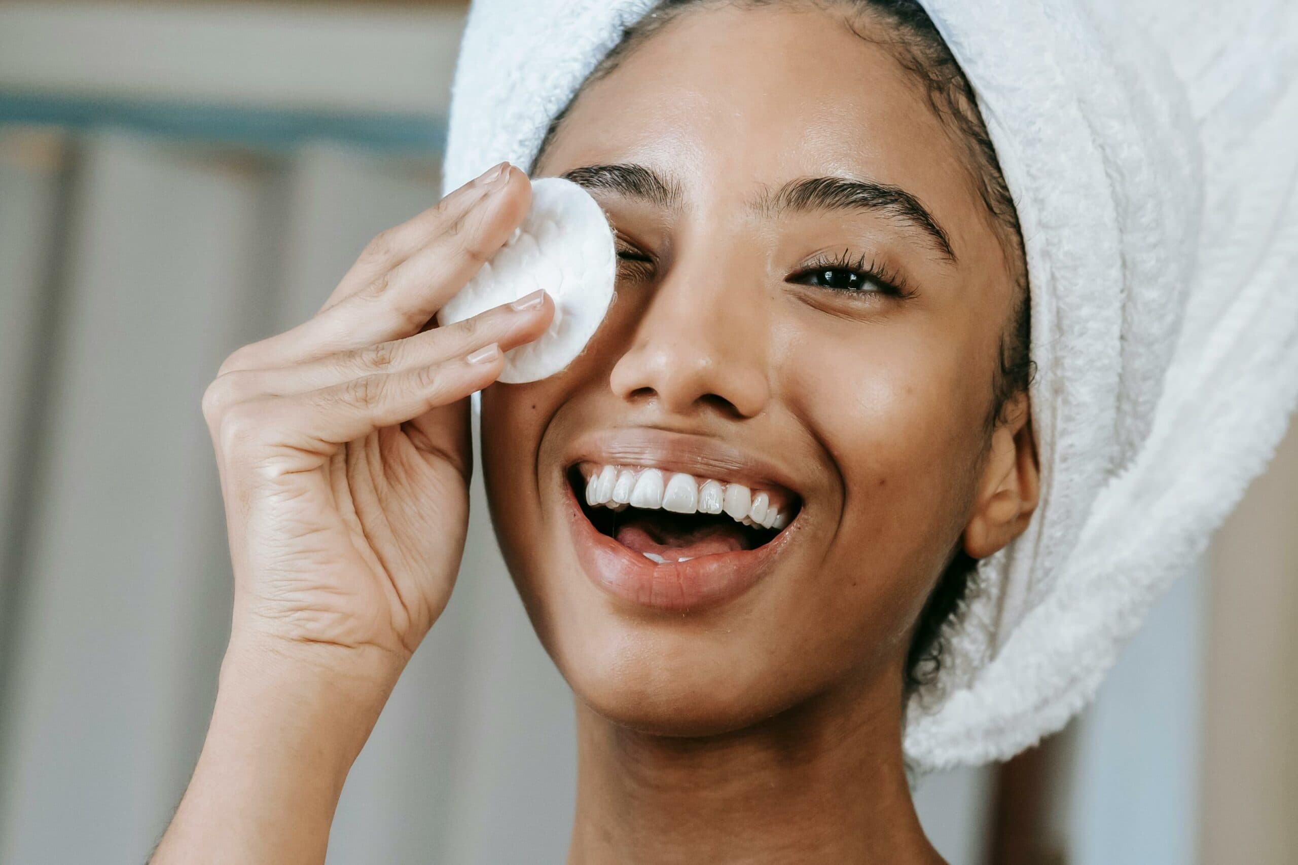 A smiling woman with a towel on her head using a cotton pad on her face, highlighting a refreshing skincare routine for healthy, glowing skin.